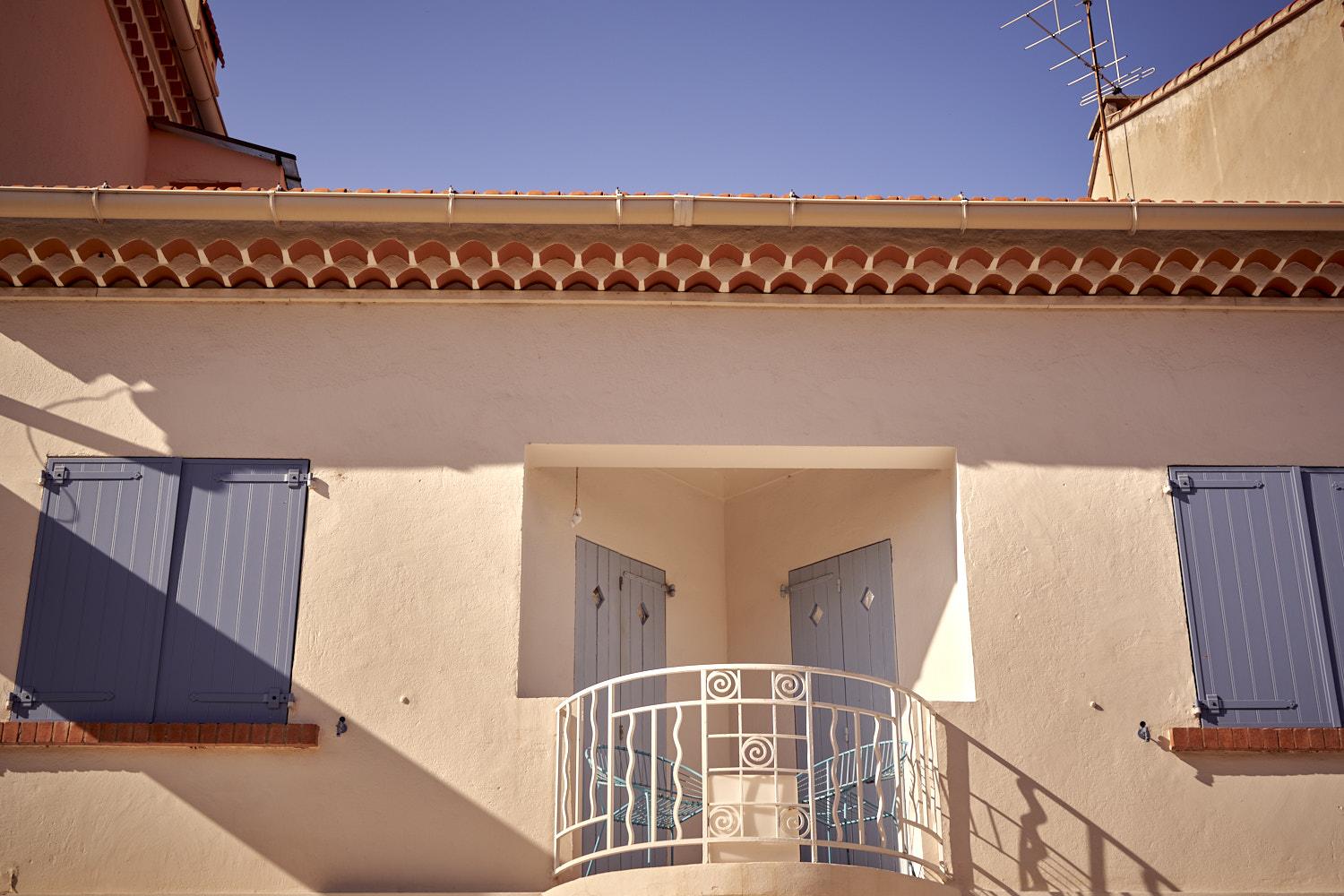 Fassade eines mediterranen Hauses mit beigem Putz, blauen Fensterl&auml;den, wei&szlig;em Balkongel&auml;nder und Ziegeldach.