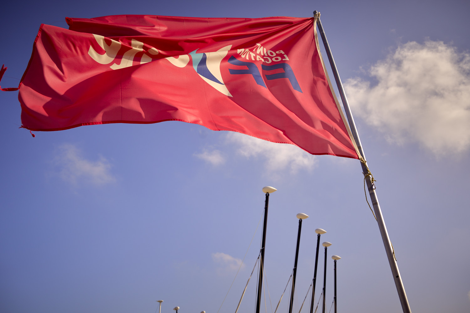 Rote Flagge mit Wappen weht an Fahnenmast gegen blauen Himmel mit wei&szlig;en Wolken.